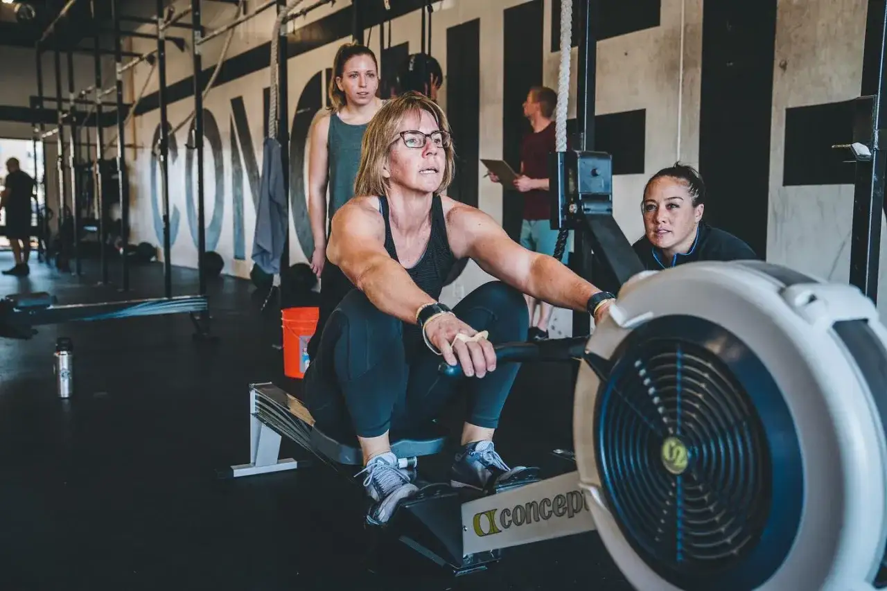 Kobieta ćwiczy na wioślarzu, co jest częścią treningu czym jest crossfit. W tle widać inne osoby i sprzęt.